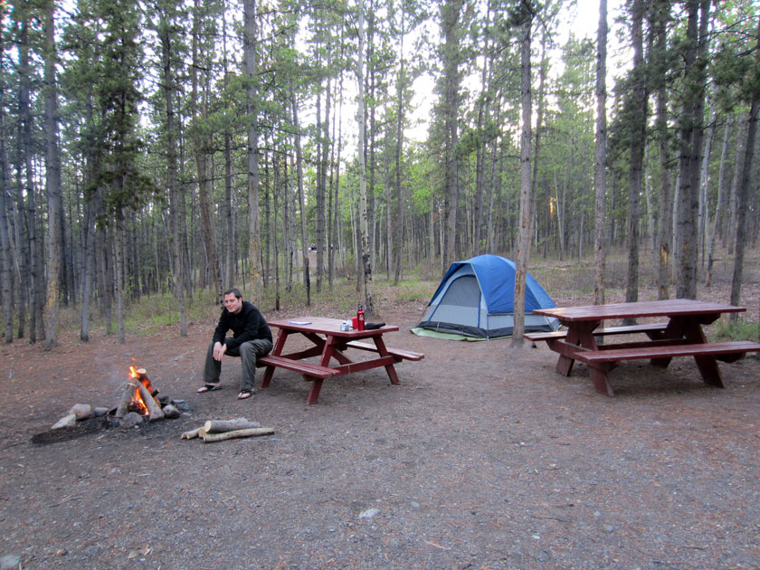 man sitting on a picnic table in front of a fire at a campsite