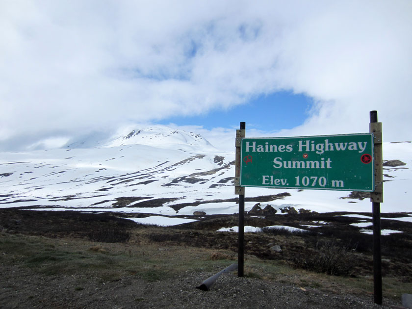 sign in front of a snowy mountain reading 'Haines Highway Summit Elev. 1070 m'